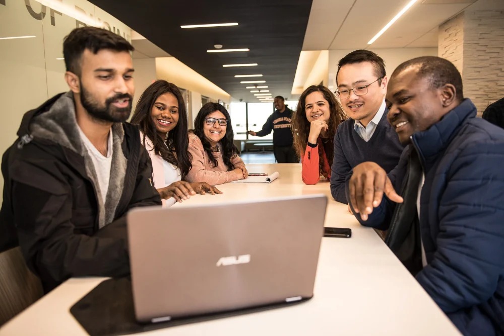 Group of UCONN MBA students sitting around a table looking at a laptop.