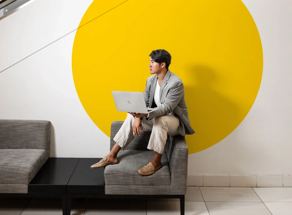 UCONN MBA student sitting on top of a couch hold a laptop in front of a wall with a large yellow circle